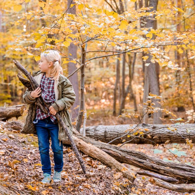Ein Mädchen steht mit einem großen Stock in der Hand im Wald und schaut zur Seite