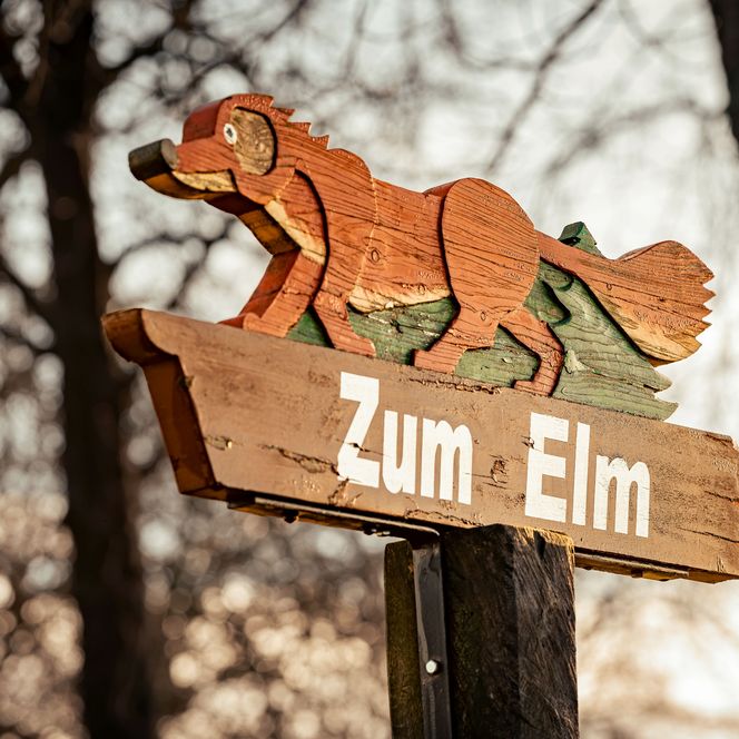 Wegweiser aus Holz mit der Aufschrift "Zum Elm" und auf dem Wegweiser befindet sich ein Fuchs aus Holz